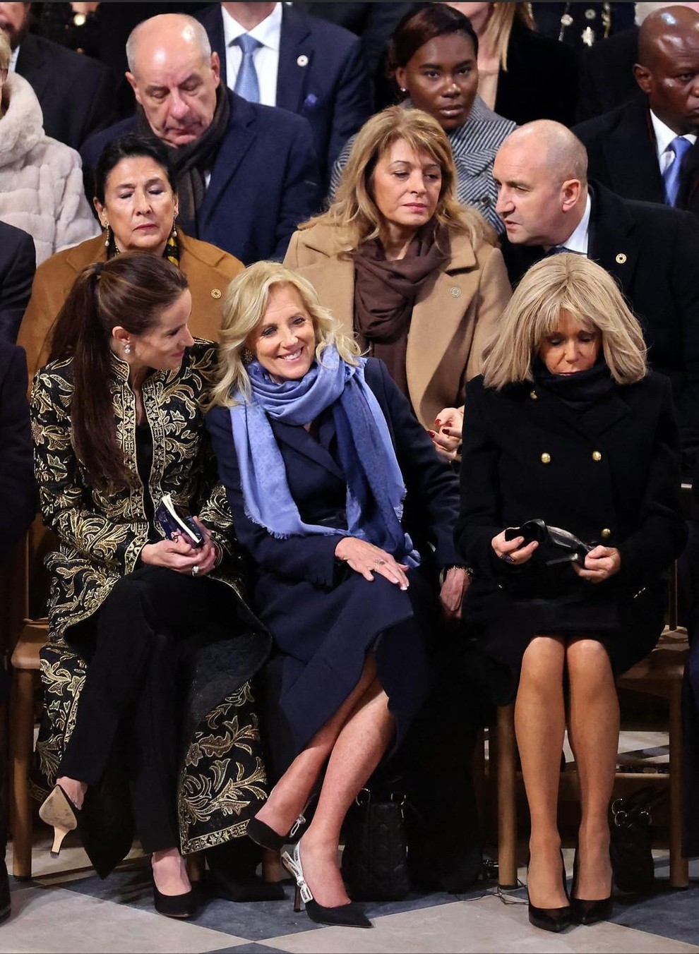 Brigitte Macron, oblečena v elegantno črno obleko s plaščem in zlatimi gumbi, je v roki nosila mini torbico znamke z …